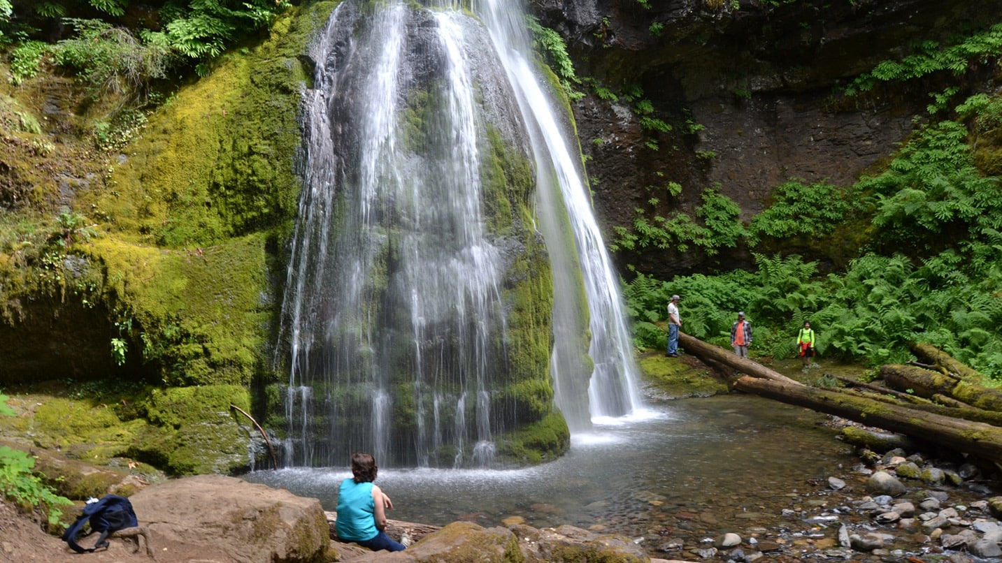 A hiker sits below the cascade of Spirit Falls, likely feeling the waterfall's mist.