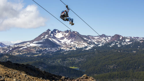 A zipliner makes a descent with a snowy mountain peak in the background.