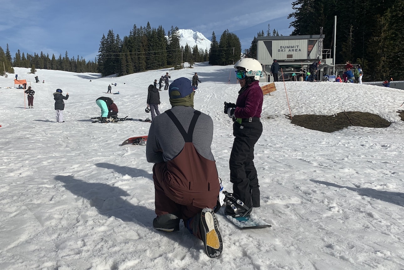A child practices snowboarding as a mentor squats next to them at Summit Ski Area.