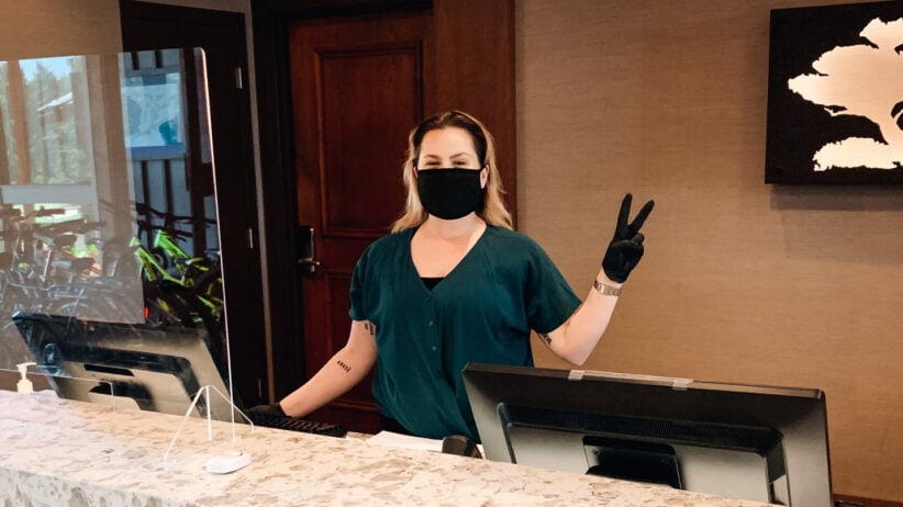 A hotel's front-desk employee wears a face covering and gloves while flashing a peace sign.