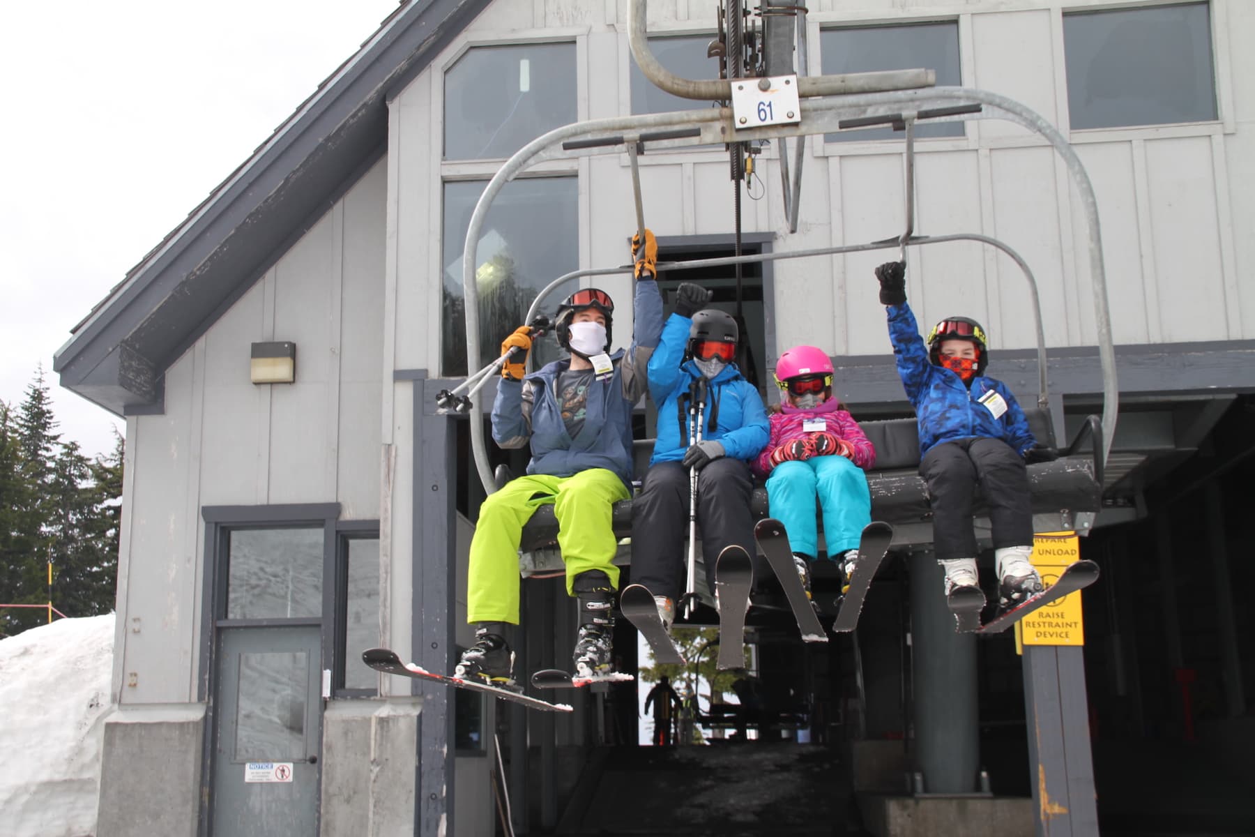 A family of four sits on a ski lift, geared up for the slopes and wearing face coverings.