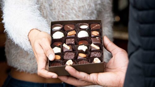 A hand passes an open box of chocolates to a friend.