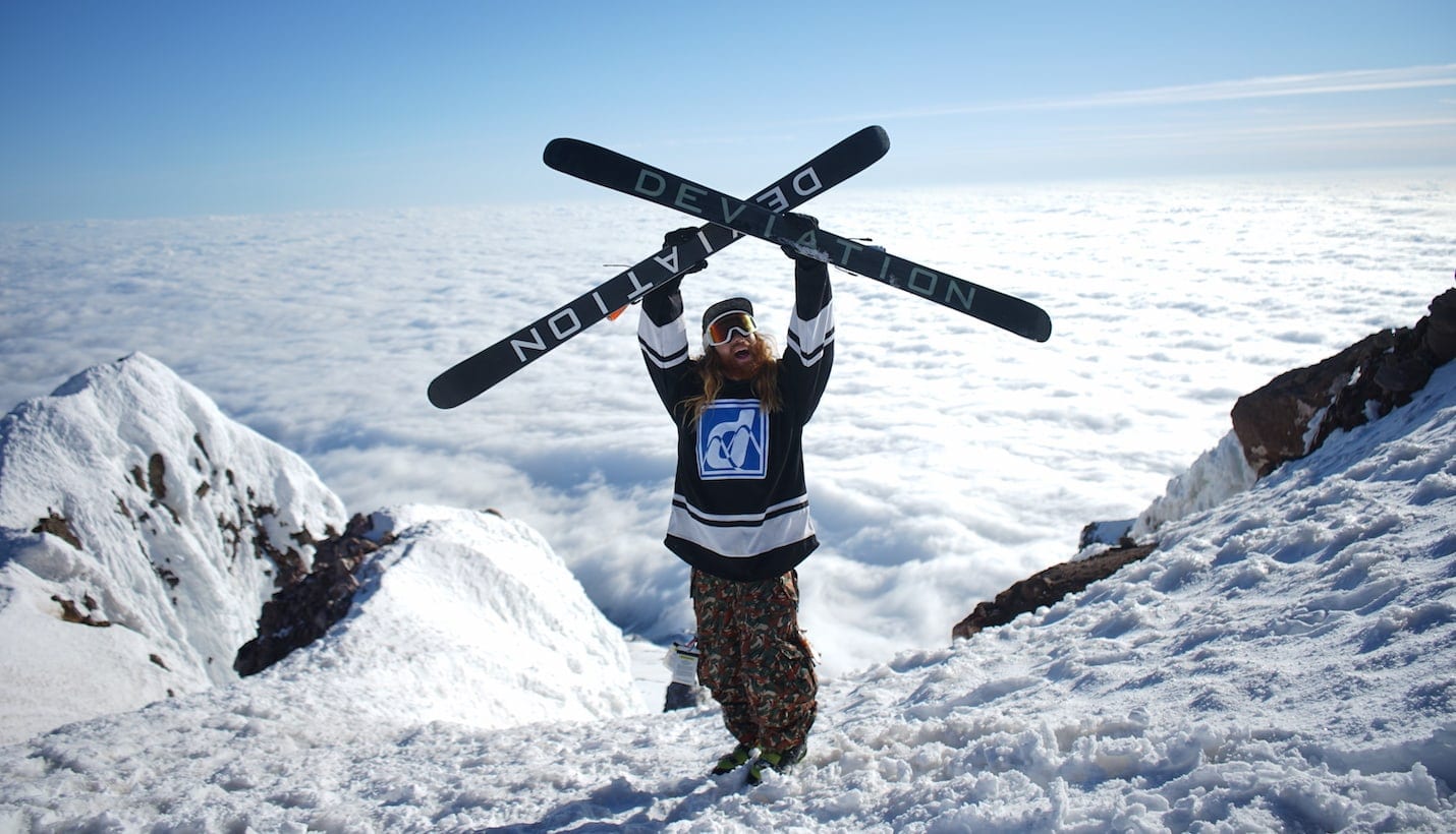 An athlete holds two skis over their head at the top of a snowy mountain.