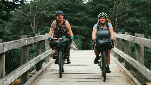 Two women bike over a wooden pedestrian bridge