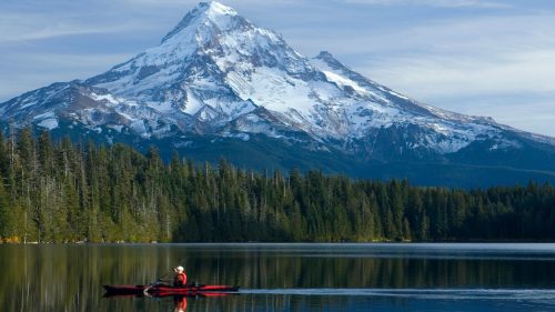 A kayaker paddles in a calm lake with a snow covered mountain in the background