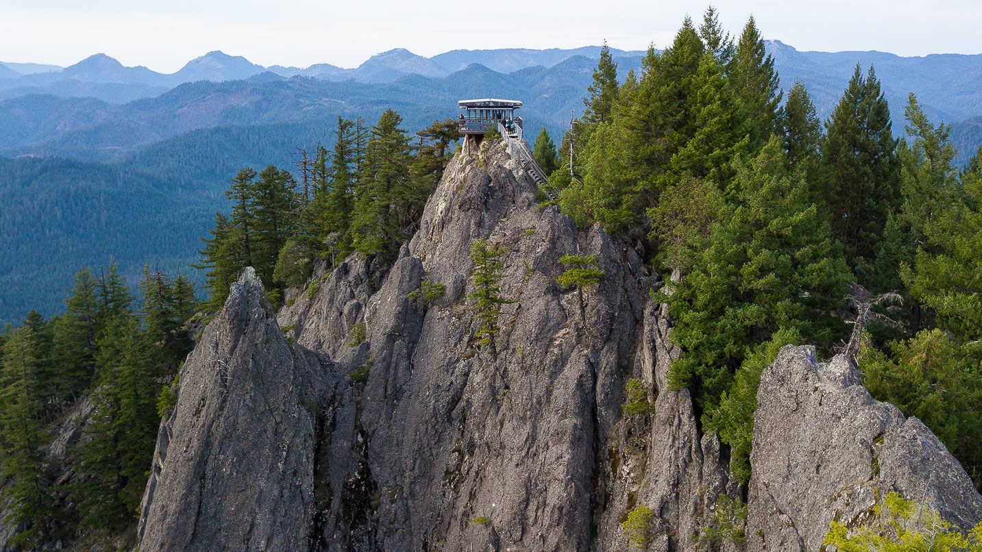 A small fire tower lookout is perched on top of rock with forested hills in the distance
