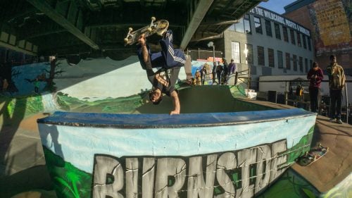 Skateboarder performs a handstand trick on edge of colorful skatepark bowl. Graffiti reads "BURNSIDE." Underpass location with onlookers and buildings in the background, urban environment.