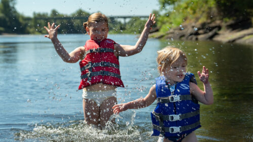 Two young children wearing lifejackets splash in a river