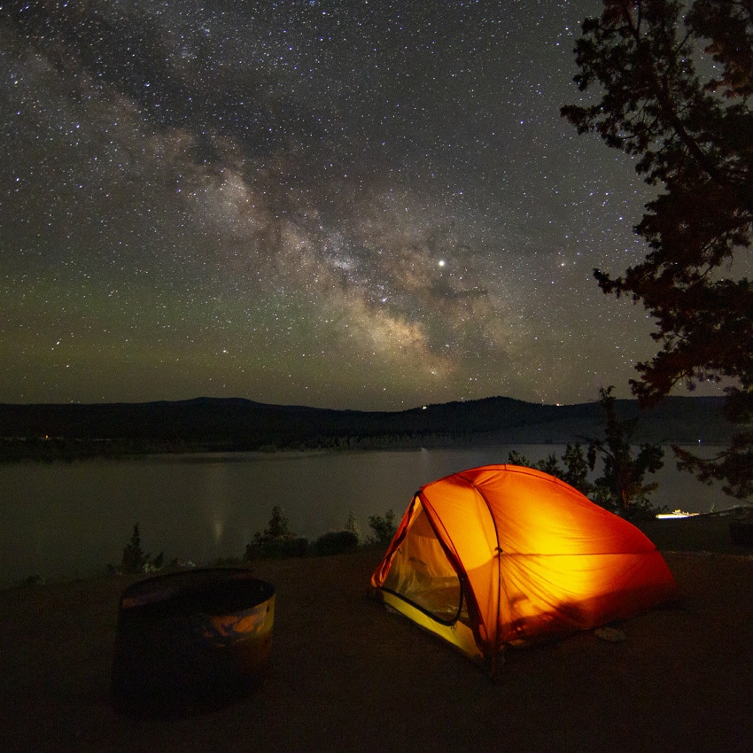A lit tent on a shore of a lake with a view of the milky way