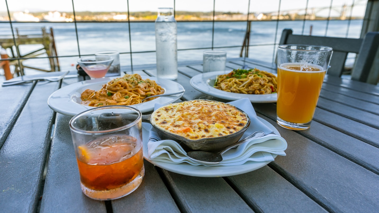 Plates of food and drinks sit on a table overlooking a bay