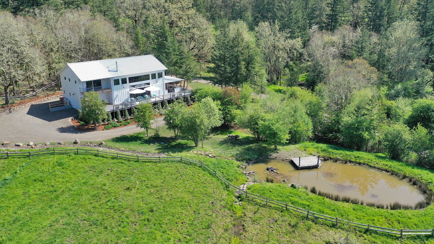 An aerial view shows a farmhouse and pond surrounded by a forest