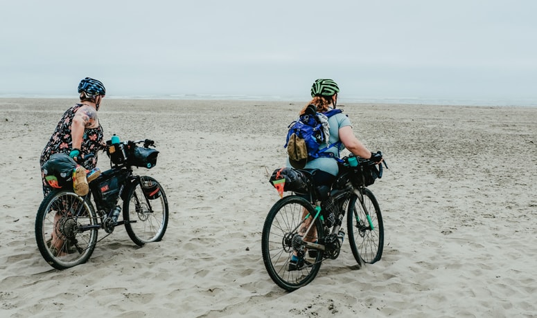 two bicyclists walking their bikes on a sandy beach