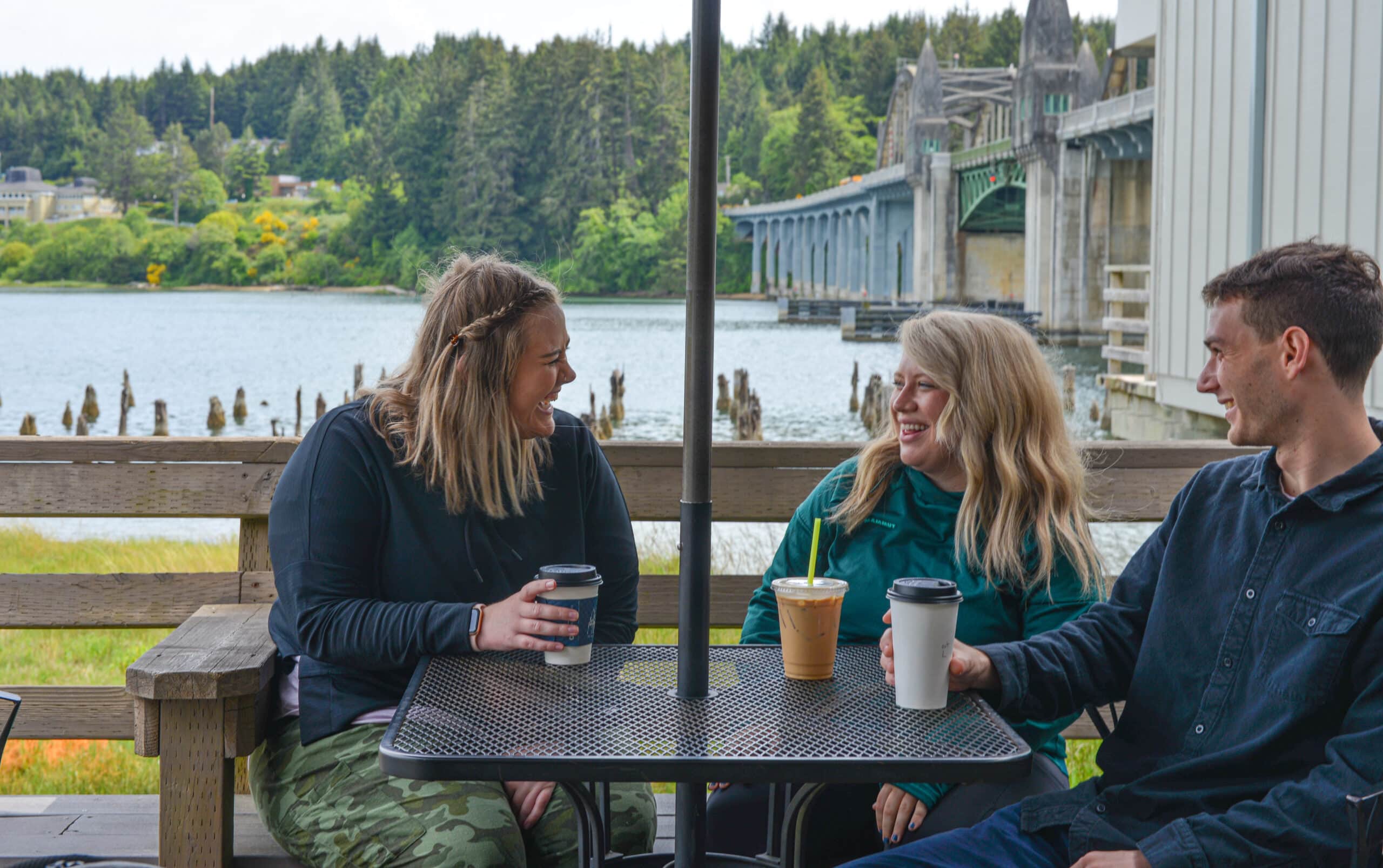 three people sit at table with coffee in front of water and bridge