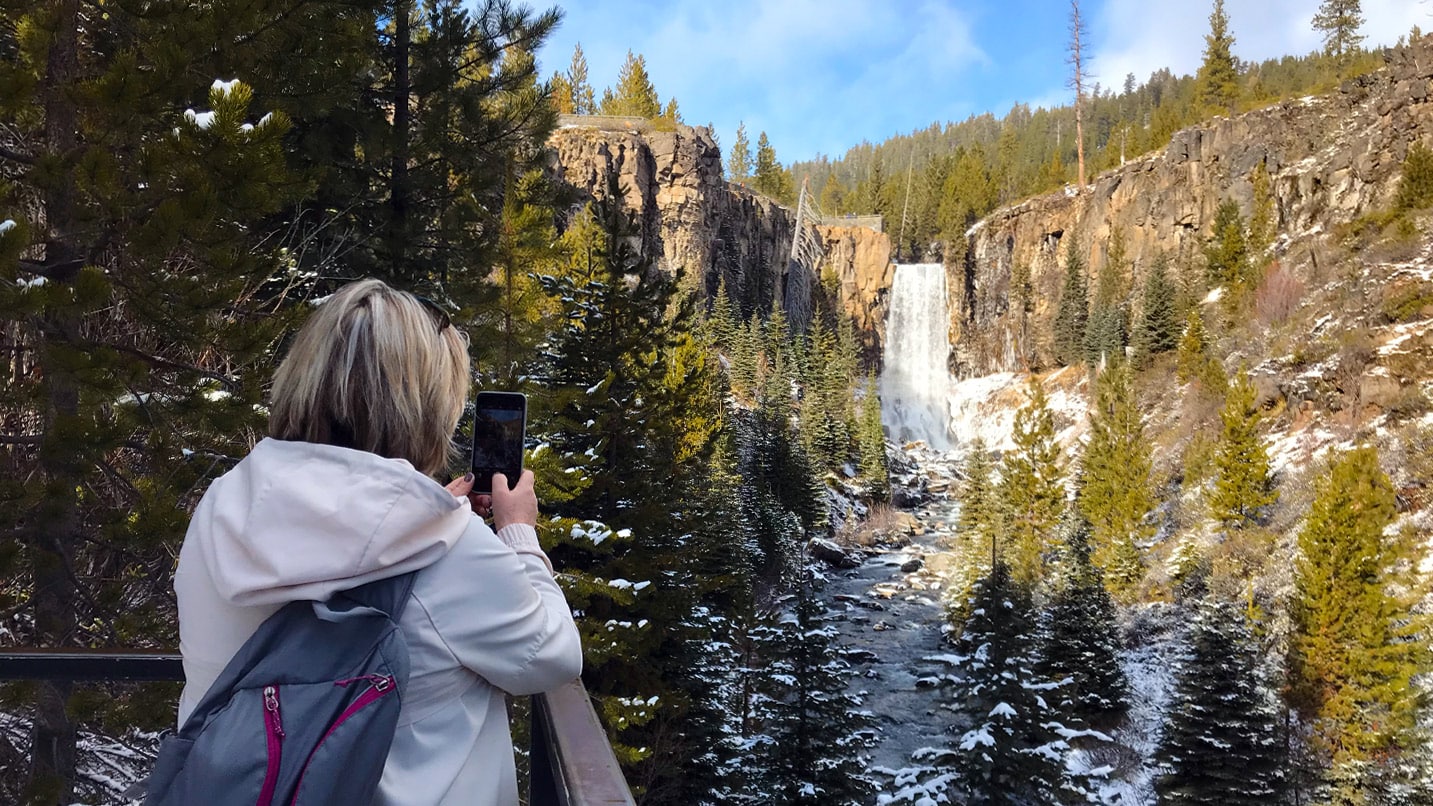 A person stands at a railing to take a photo of a snowy waterfall