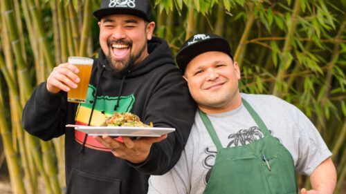 A man holds a plate of food and a beer and another man leans in