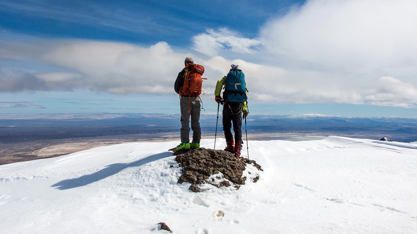 Two people stand on a snowy mountain looking at blue sky and clouds