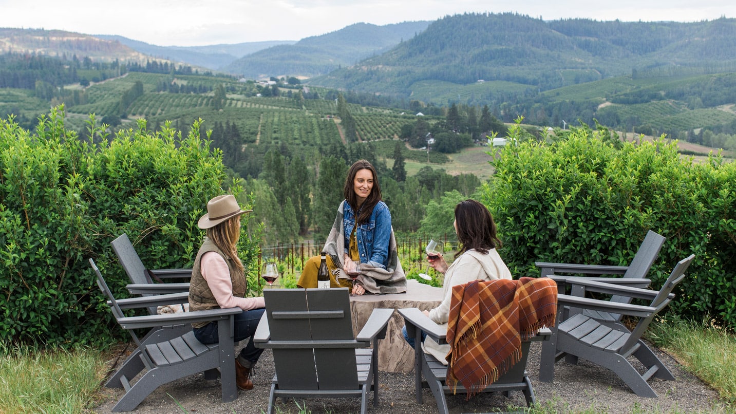 Three women sit around a table with wine in front of a panoramic valley