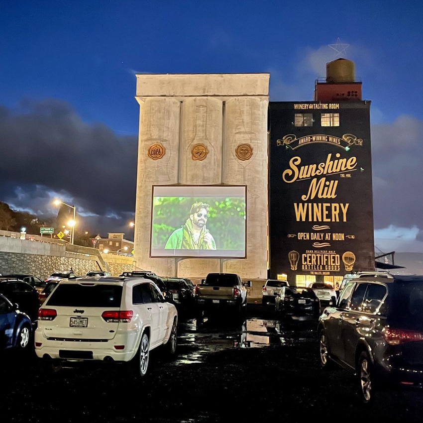 A movie plays on an outdoor silo in front of parked cars