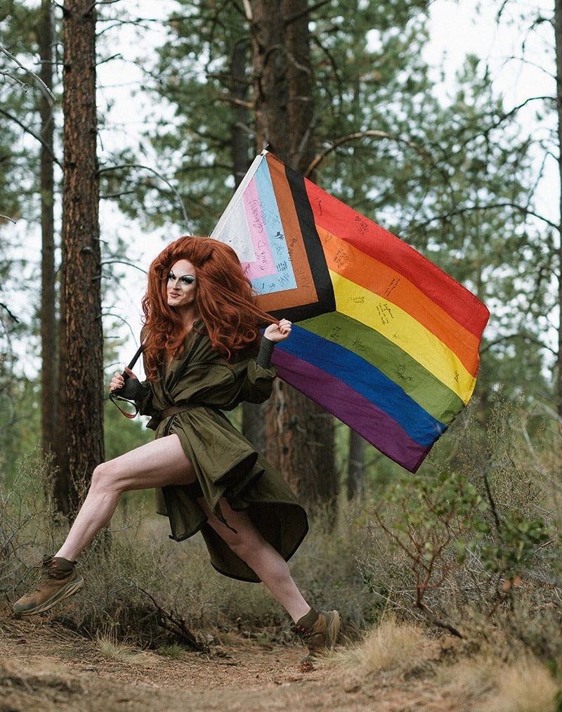 person wearing green coat in forest with large rainbow flag