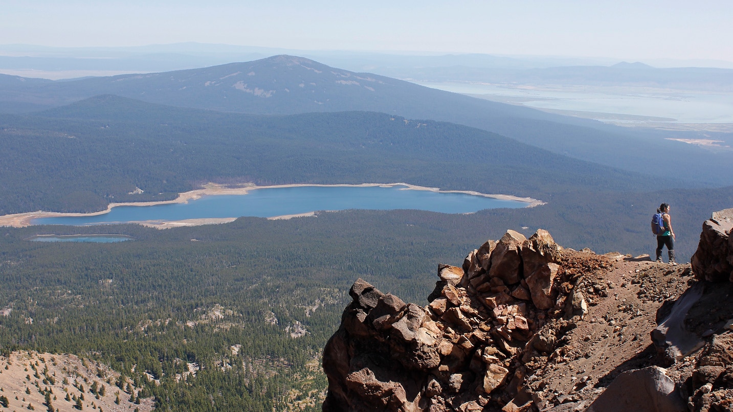 A person stands on top of a rocky mountain overlooking a lake and valley below