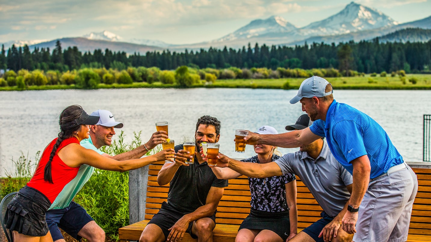 A group of people cheers with glasses of beer at an edge of a lake with snow-capped peaks in the background