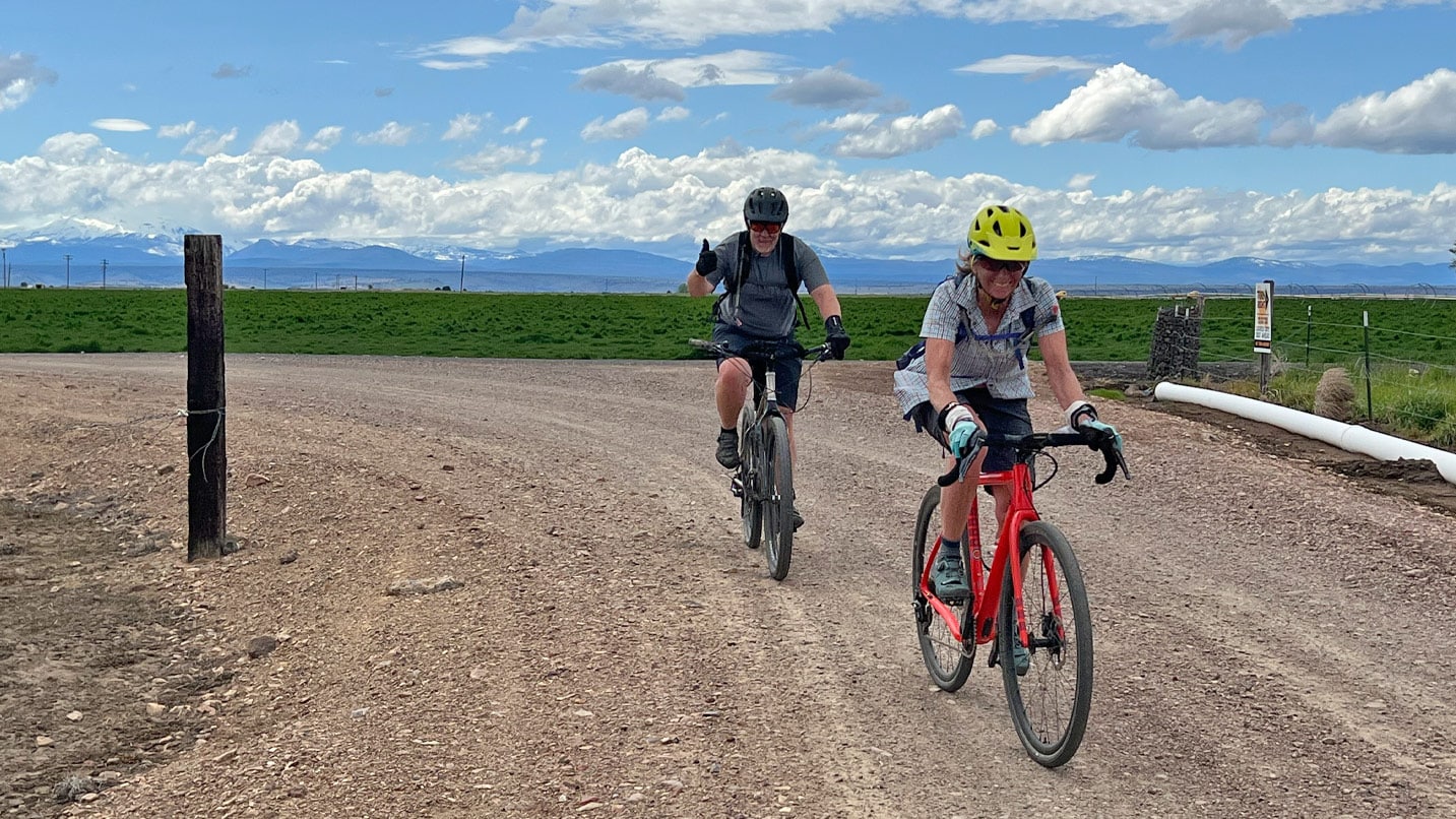two cyclists pedal on gravel road with clouds and blue sky in background