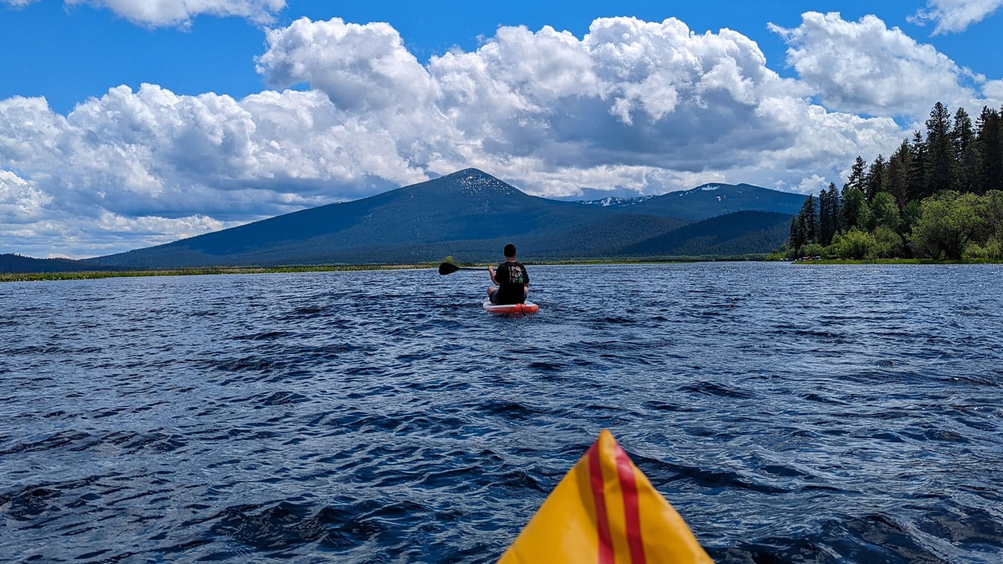 person sits on paddleboard in water with mountain and clouds in distance
