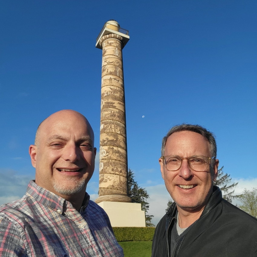 two men stand outside tall column with blue sky