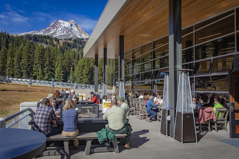people dining on outdoor patio with snow-capped mountain and trees in distance