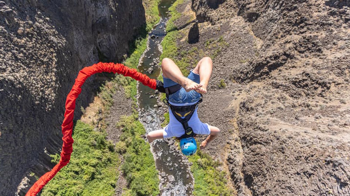 person hangs upside down over river, attached by red bungee cord