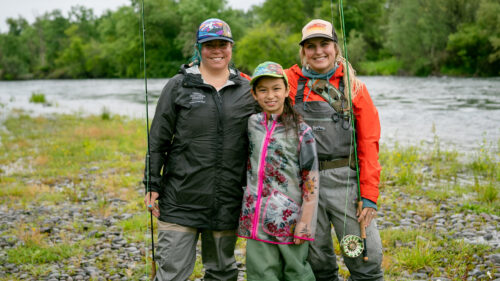 Two women and a young girl stand on the edge of a river holding fishing rods