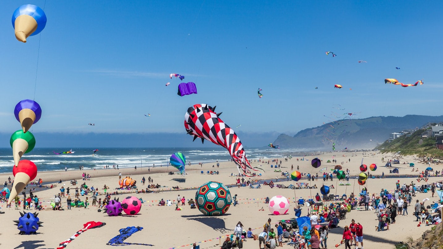 Many colorful kites fly above a sandy beach