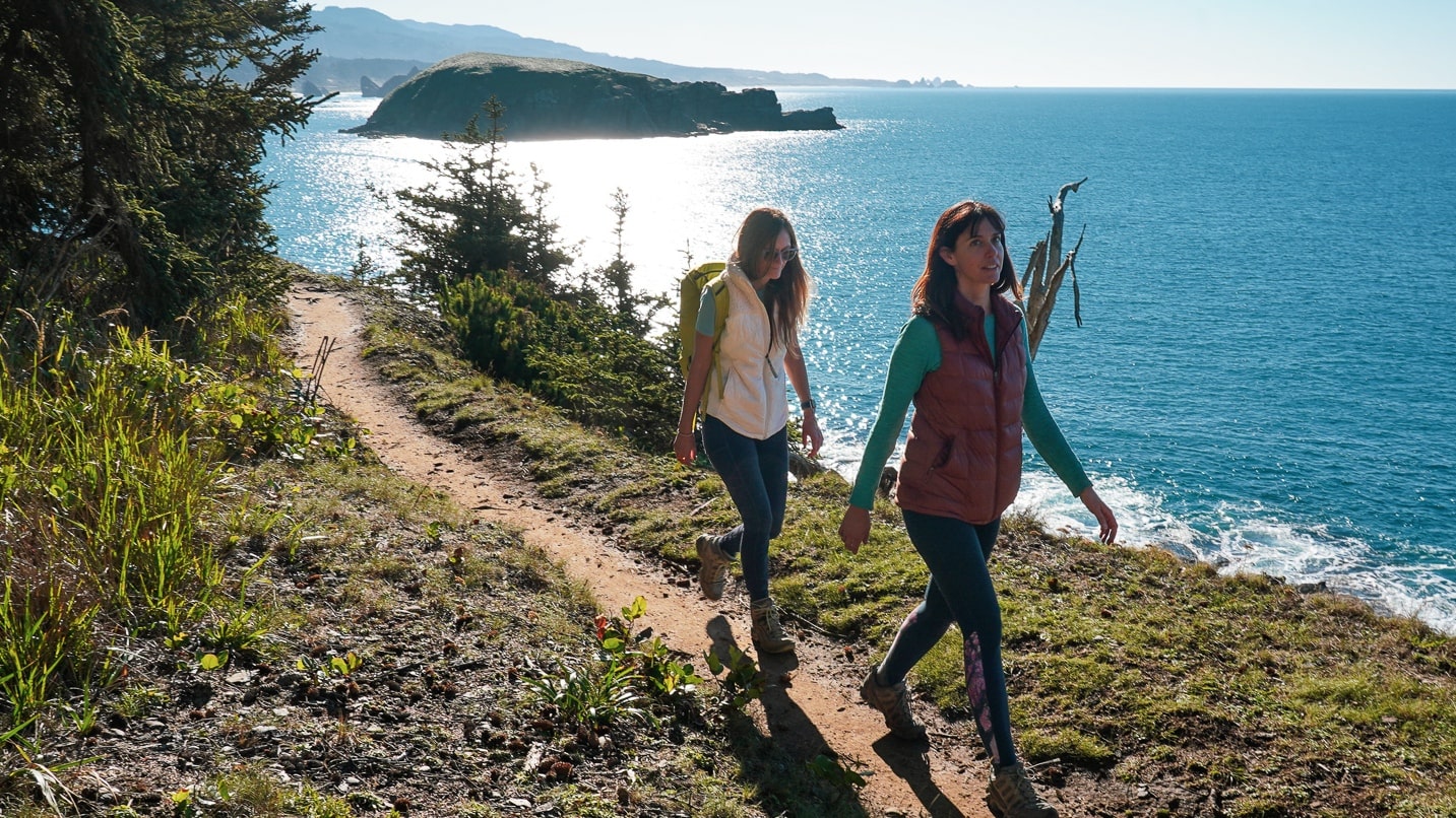 two women hike on a trail with ocean in background