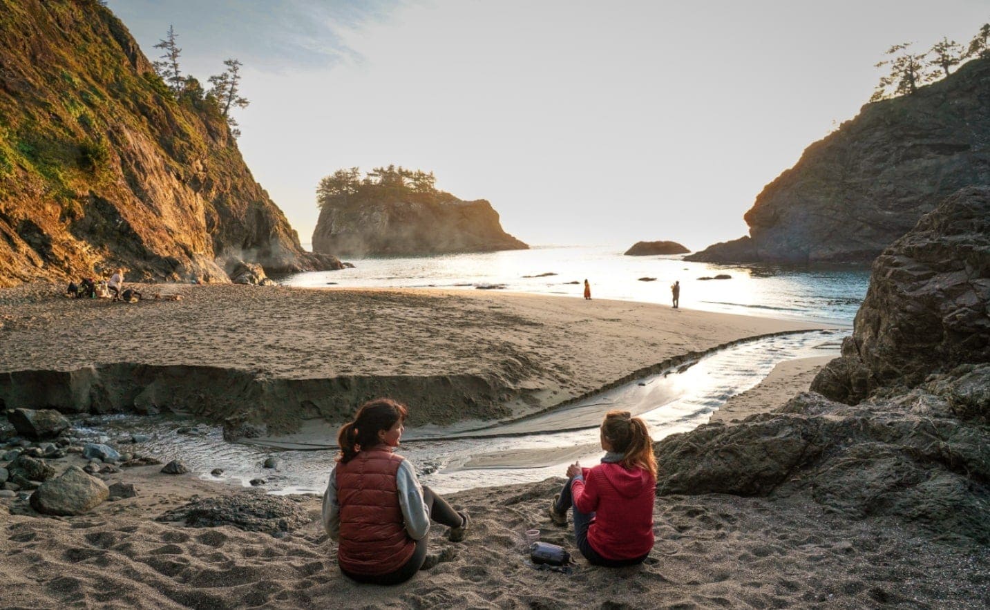 two women sit next on sand looking out to ocean and sea stacks