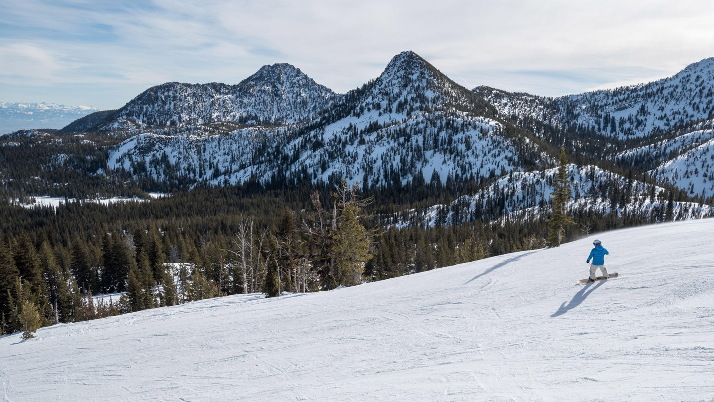 Snowy mountain ranges and pine trees in the background. In the foreground, we see an aerial view of a snowboarder going down an incline.