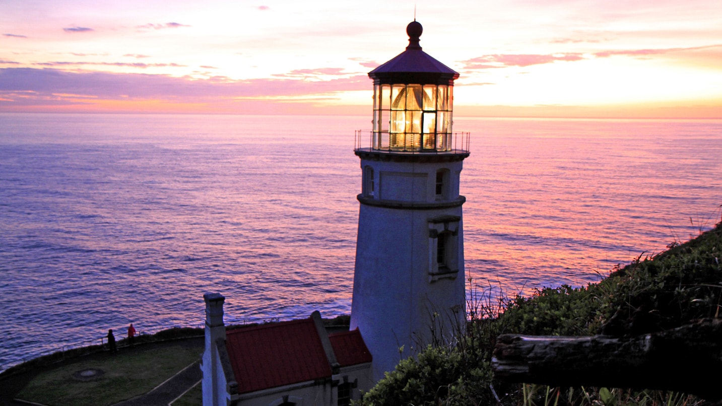 A sunset over a lighthouse. The ocean water appears of a mix of pink and purple like the sky.