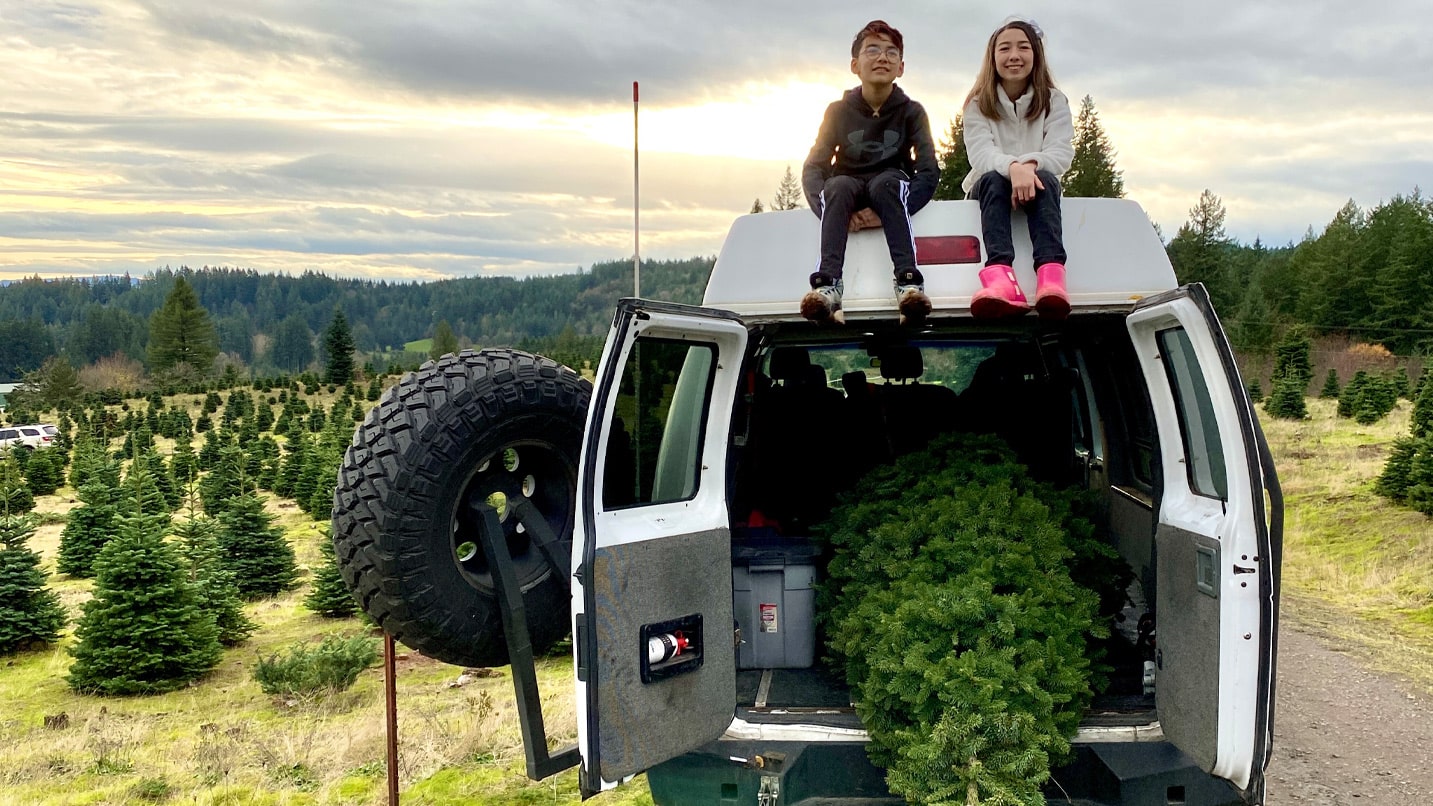 Two children sit smiling atop a white van that has it's back doors open. Inside the van is a small pine tree.