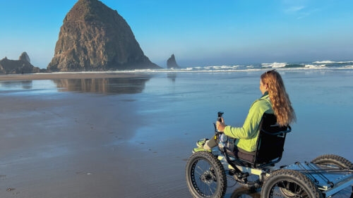A woman in a power wheelchair on a beach. In the background a large haystack rock towers over the shore.