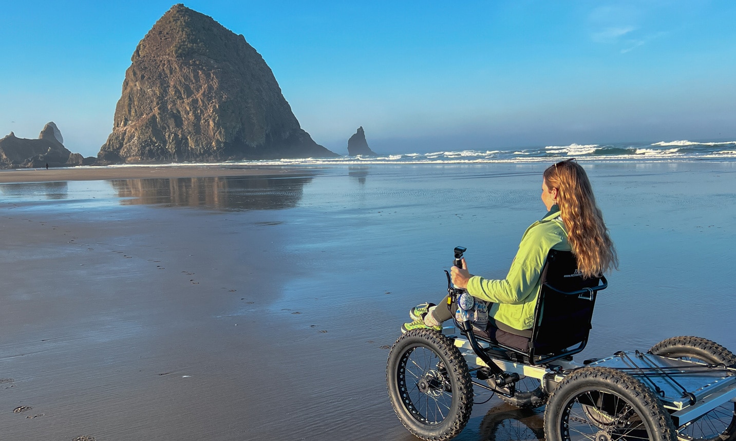 A woman in a power wheelchair on a beach. In the background a large haystack rock towers over the shore.