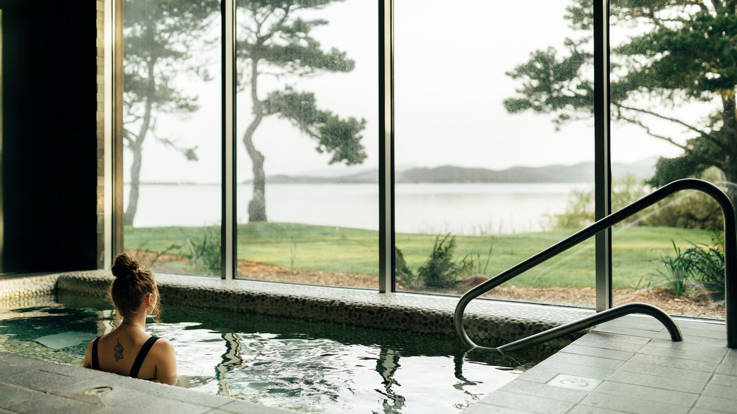 A person sits in a hot tub with views of the ocean through large windows