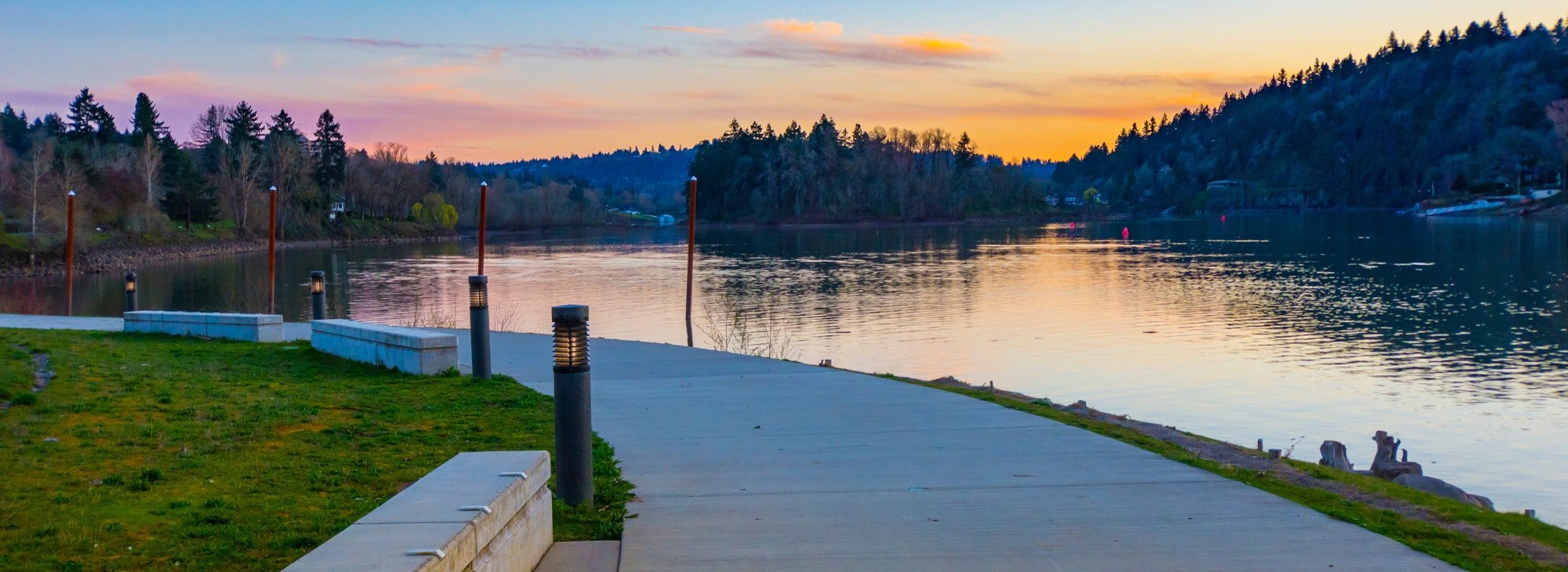 A plank path curves along a river. The sunset colors the sky hues of pink and orange and blue.