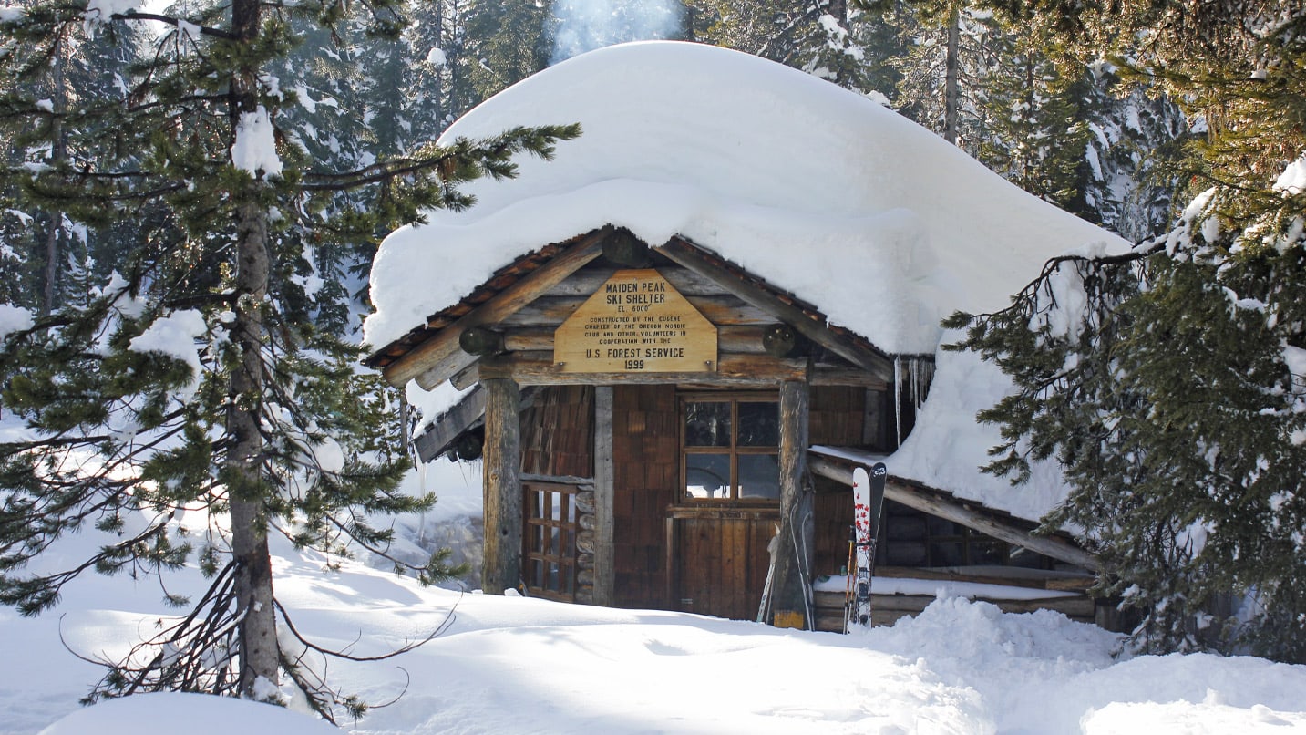 View of a small wooden cabin in the woods. Snow is piled high on its roof and surrounding area.