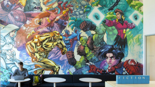 An art mural of superheroes along the wall of the Milwaukie Library. A person on a laptop sits in front of the mural at a table.