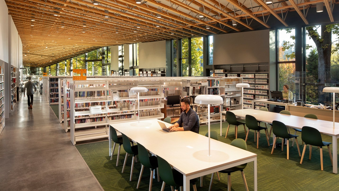 Inside of the Milwaukie Library. A man on a laptop sits at a large table. A person walks between shelves of books.