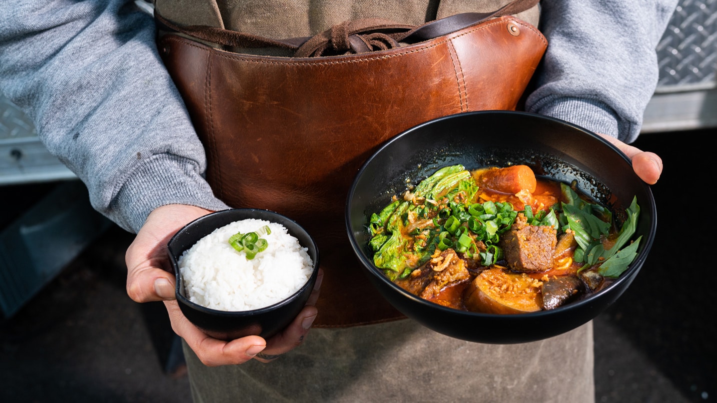 Lower body shot of a person holding a small bowl of rice and a larger bowl of food with pork and vegetables.