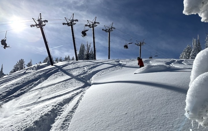 A snowboarder riding through deep powder under the chairlift. 