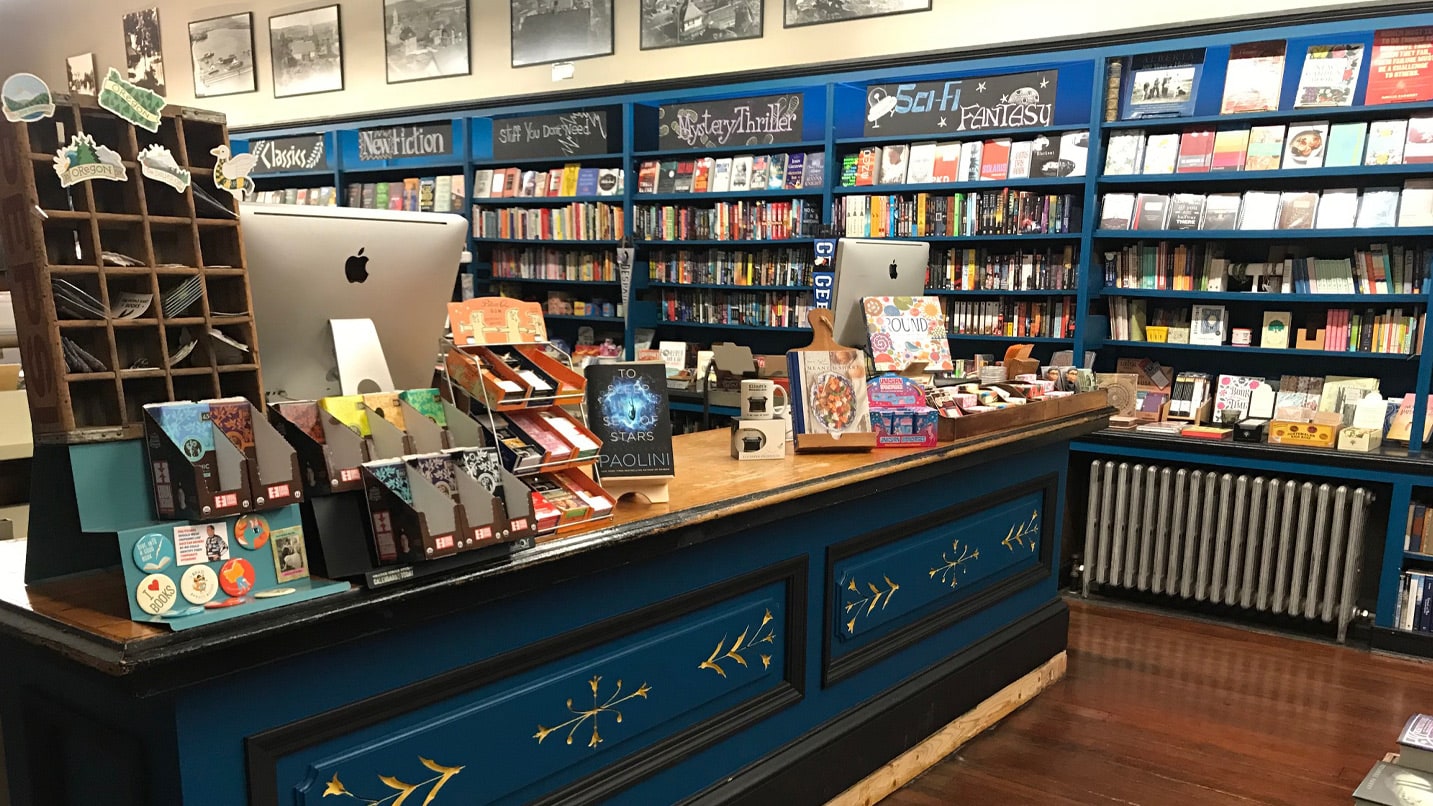 Inside of a shop. Shelves are filled with books on display. The cashier counter is painted a deep blue with gold accents.