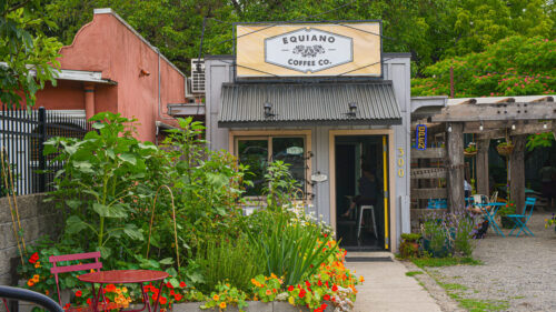 Exterior shot of a small coffee shop. Plants surround a small seating area.