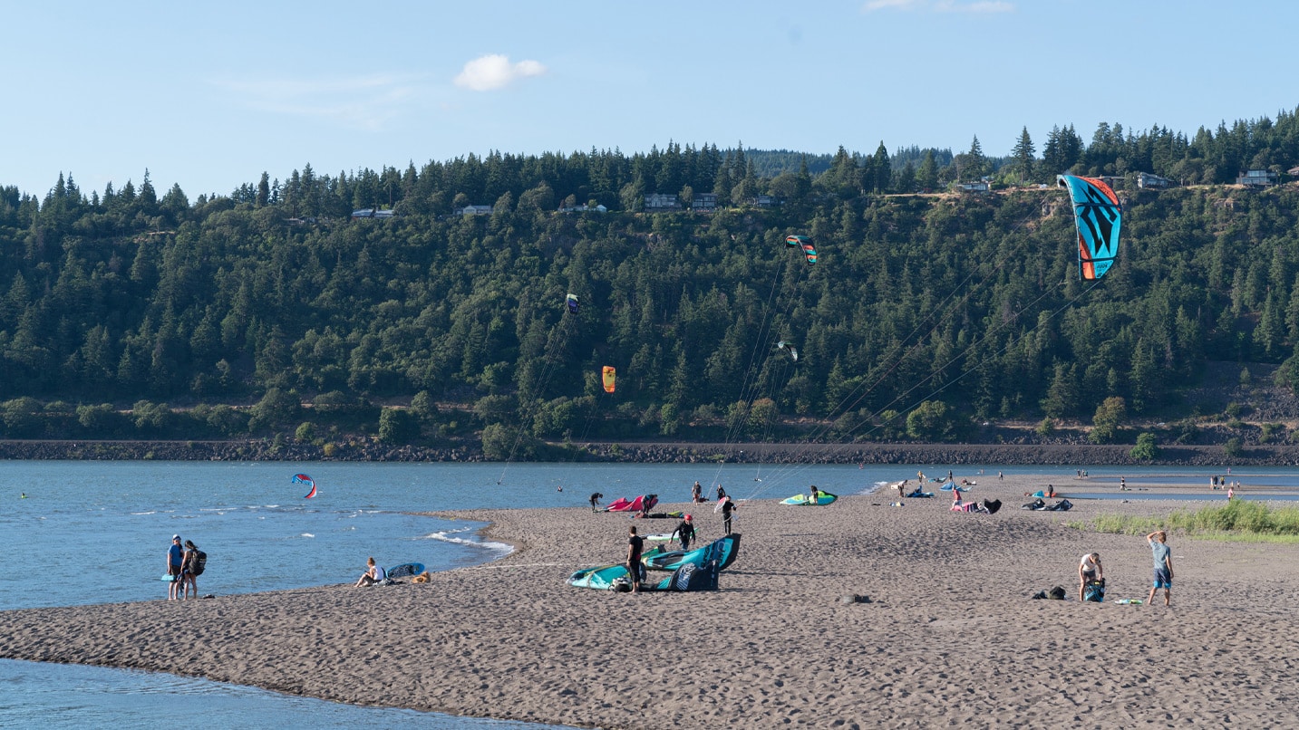 A sandy beach area along the Columbia river. People lounge, swim, and fly kites.