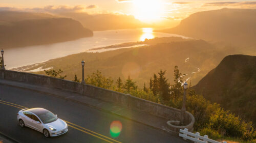An electric vehicle drives along a byway against a backdrop of the sun setting over the Columbia River Gorge.